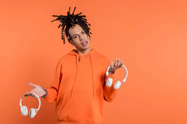 Uncertain multiracial man with dreadlocks holding different wireless headphones and looking at camera on coral background — Stock Photo