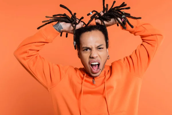 Tattooed and emotional multiracial man standing with opened mouth and pulling dreadlocks on coral background — Stock Photo