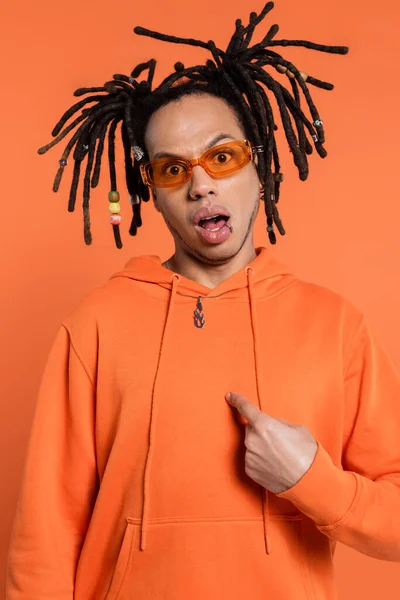 Shocked multiracial man in hoodie and stylish sunglasses pointing at himself isolated on coral — Stock Photo