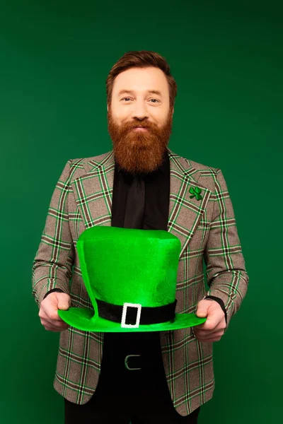 Smiling bearded man in jacket holding hat while celebrating saint patrick day isolated on green — Stock Photo