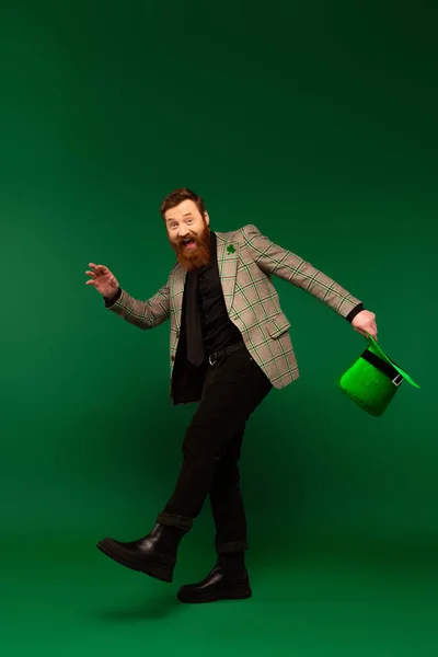 Full length of cheerful bearded man holding hat during saint patrick day on green background — Stockfoto