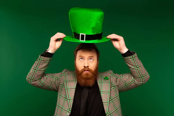 Бородатий чоловік в куртці дивиться на капелюх під час святкування святого патруля ізольовано на зеленому — стокове фото
