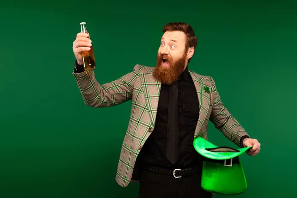 Excited bearded man looking at bottle of beer and hat during saint patrick day isolated on green - foto de stock