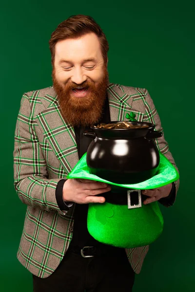 Cheerful bearded man holding pot with coins and hat during saint patrick day isolated on green — Stockfoto