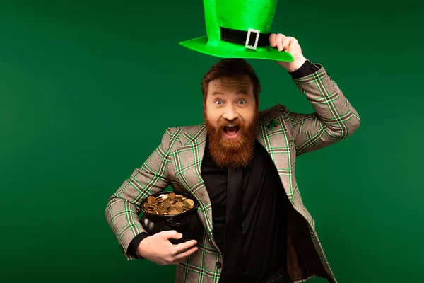 Волнующий бородатый держа шляпу и горшок с монетами во время святого Патрика день изолирован на зеленый — стоковое фото