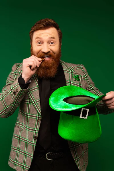 Bearded man biting coin and holding hat during saint patrick day isolated on green — Stock Photo