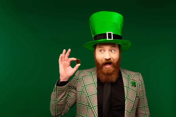 Amazed bearded man in hat and blazer with clover holding coin isolated on green — Stock Photo