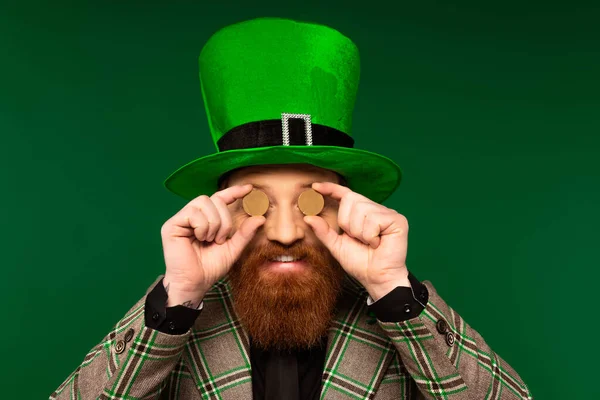 Smiling bearded man in hat holding coins near eyes isolated on green — Stock Photo