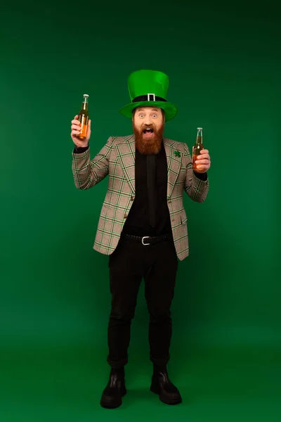 Comprimento total de homem surpreso em chapéu segurando garrafas de cerveja no fundo verde — Fotografia de Stock