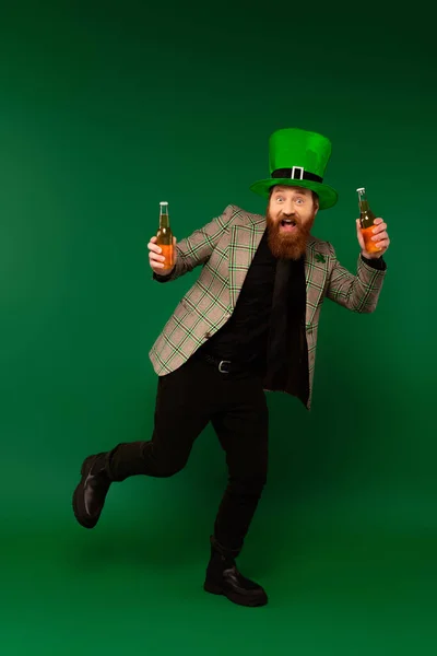 Повна довжина збудженого чоловіка в капелюсі, що святкує день святого патруля і тримає пляшки пива на зеленому фоні — стокове фото