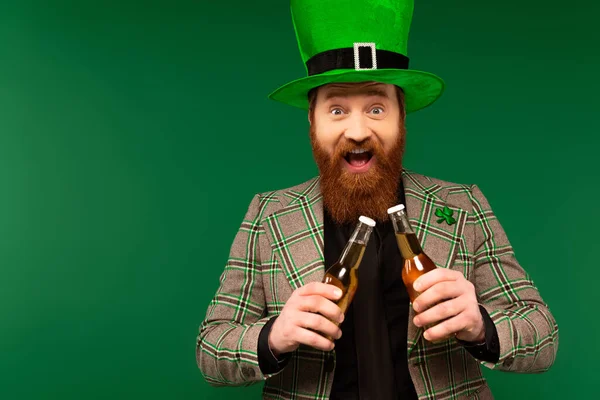 Excited bearded man in hat holding beer bottles during saint patrick day isolated on green - foto de stock