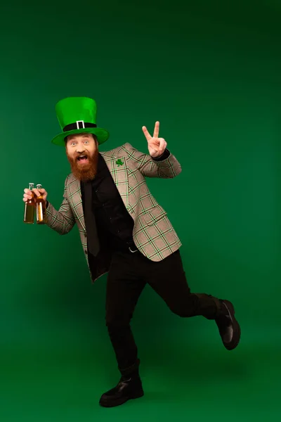 Emocionado hombre barbudo con sombrero sosteniendo botellas de cerveza y mostrando signo de paz sobre fondo verde - foto de stock