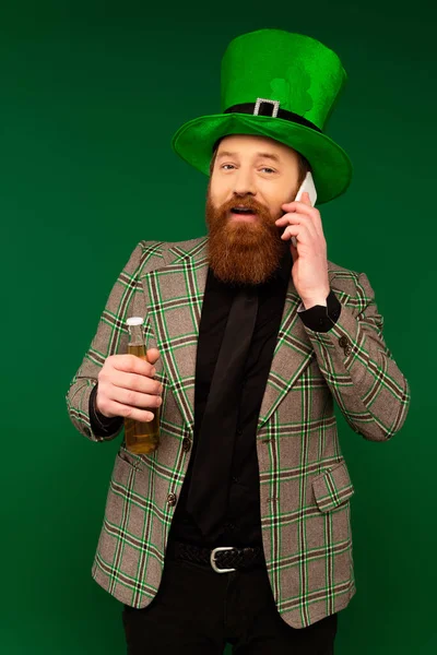 Бородатый мужчина в шляпе разговаривает на смартфоне и держит бутылку пива изолированы на зеленый — стоковое фото