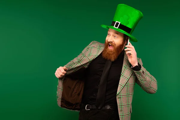 Excited bearded man in jacket talking on smartphone during saint patrick day isolated on green — Photo de stock