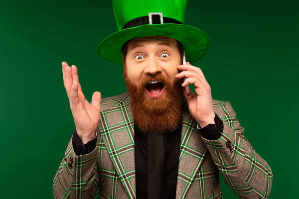 Uomo barbuto sbalordito in cappello parlando su smartphone durante il santo patrick day isolato sul verde — Foto stock