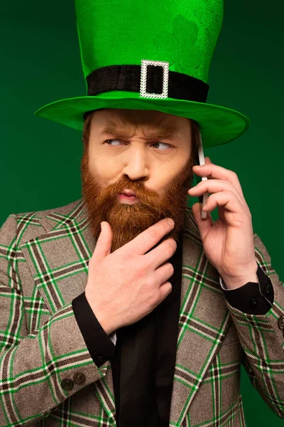 Задумчивый бородатый мужчина в шляпе с клевером разговаривает на смартфоне изолированы на зеленый — стоковое фото