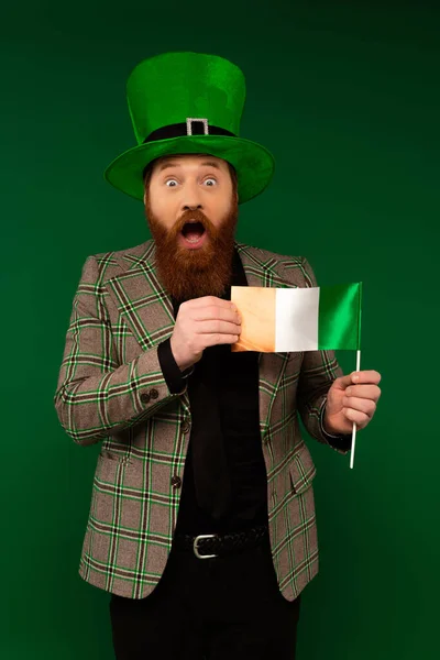 Шокированный бородатый мужчина в шляпе, держащий ирландский флаг изолированным на зеленом — стоковое фото