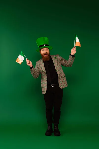 Full length of cheerful bearded man in hat holding Irish flags on green background — Stockfoto