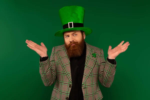 Confused bearded man in hat with clover doing shrug gesture isolated on green — Foto stock
