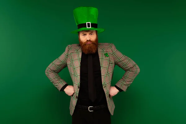 Злой бородатый мужчина в шляпе с клевером, держащимся за руки на бедрах, изолированных на зеленом — стоковое фото