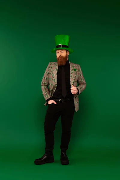 Longitud completa del hombre barbudo en trébol en la chaqueta y en el sombrero posando sobre fondo verde - foto de stock