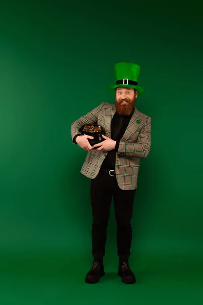 Веселый бородатый мужчина в шляпе держит горшок с золотыми монетами на зеленом фоне — стоковое фото