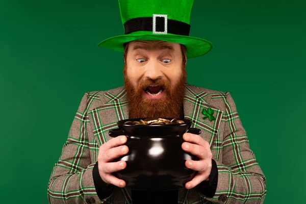 Hombre sorprendido en sombrero sosteniendo olla con monedas de oro aisladas en verde - foto de stock