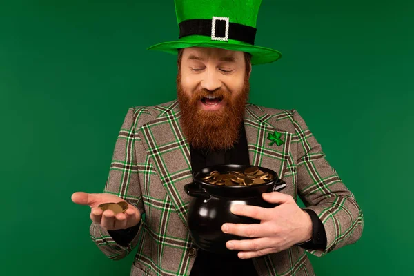 Cheerful man in hat holding golden coins and pot during saint patrick day isolated on green — Photo de stock