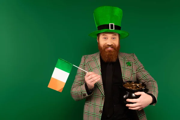 Uomo positivo in cappello con trifoglio con bandiera irlandese e pentola con monete isolate sul verde — Foto stock