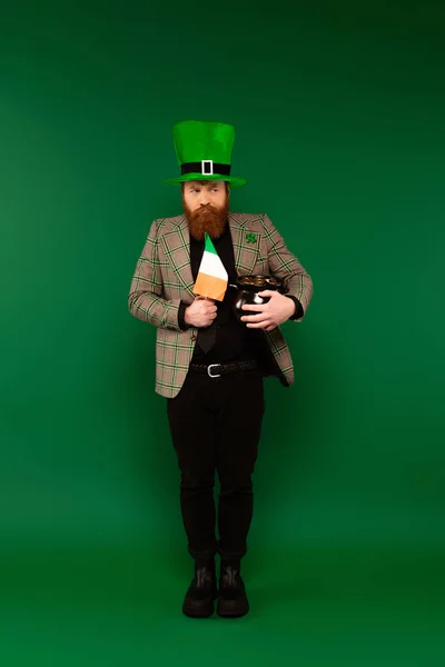 Hombre pensativo con sombrero sosteniendo bandera irlandesa y bote con monedas sobre fondo verde - foto de stock
