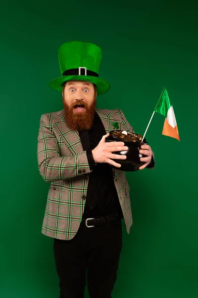 Homem barbudo chocado em chapéu segurando panela com moedas e bandeira irlandesa isolada em verde — Fotografia de Stock