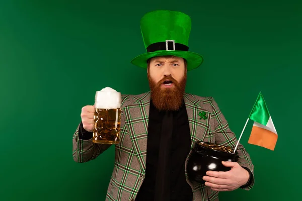 Hombre barbudo con sombrero sosteniendo cerveza y olla con monedas y bandera irlandesa aislada en verde - foto de stock