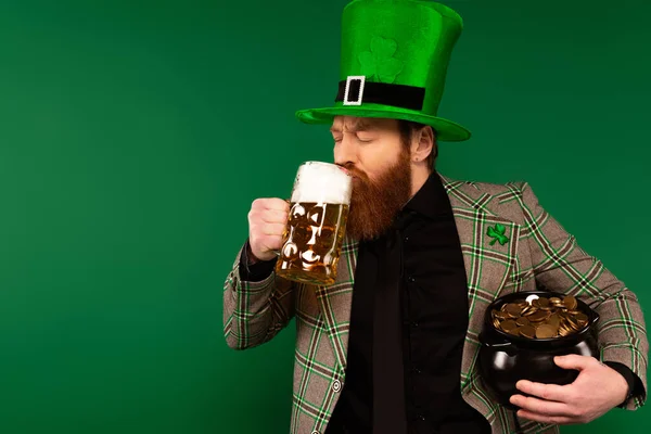 Hombre barbudo en sombrero bebiendo cerveza y sosteniendo la olla con monedas aisladas en verde - foto de stock