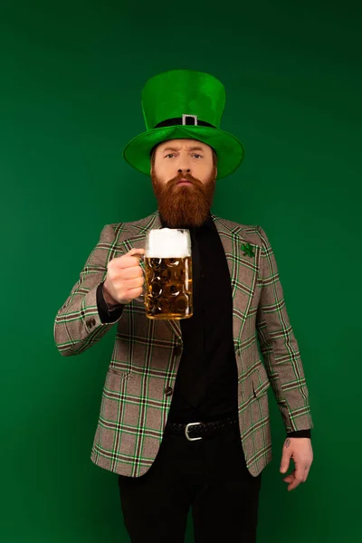 Homem barbudo sério de chapéu com trevo segurando copo de cerveja isolado em verde — Fotografia de Stock