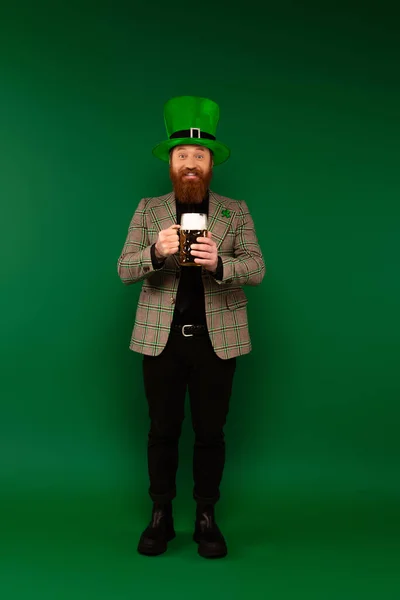Pleine longueur d'homme barbu souriant dans un chapeau tenant un verre de bière sur fond vert — Photo de stock