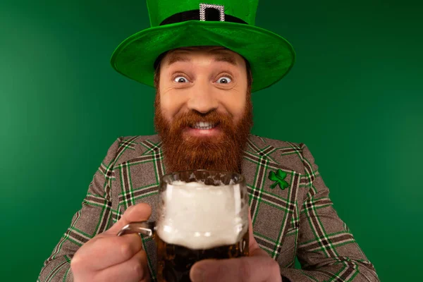 Allegro uomo barbuto in cappello che tiene la birra e guarda la macchina fotografica durante il giorno santo Patrick isolato sul verde — Foto stock