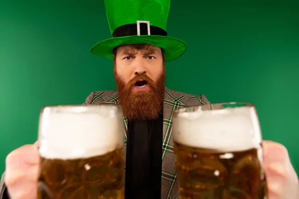 Homem barbudo sério em chapéu segurando copos de cerveja isolados em verde — Fotografia de Stock