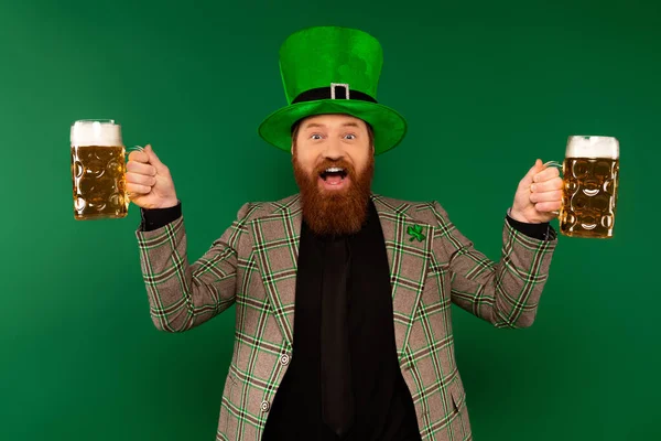 Homem barbudo positivo em chapéu celebrando dia santo patrick e segurando copos de cerveja isolado em verde — Fotografia de Stock