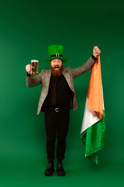 Excited bearded man in hat holding glass of beer and Irish flag on green background — Stock Photo
