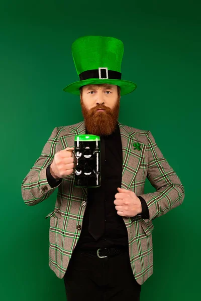 Homme barbu sérieux dans le chapeau tenant verre de bière isolé sur vert — Photo de stock