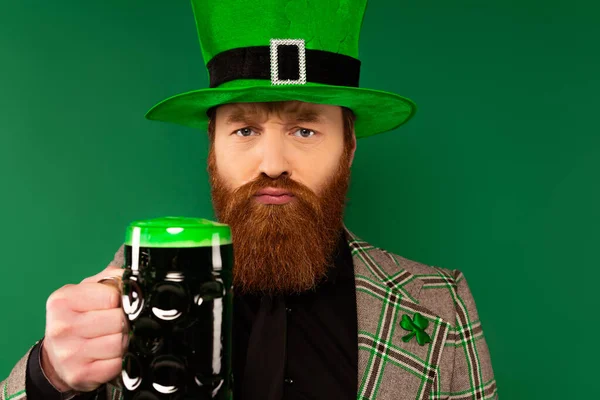 Sérieux barbu homme dans chapeau tenant verre de bière et regardant caméra isolé sur vert — Photo de stock