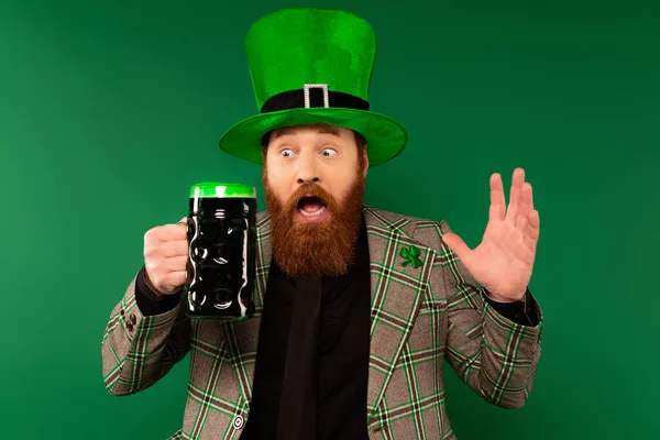 Удивительный бородатый мужчина в шляпе с клевером, смотрящий на стакан пива, изолированного на зеленом — стоковое фото