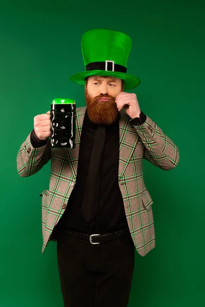 Pensive man in hat with clover looking at beer and touching moustache isolated on green — Stockfoto