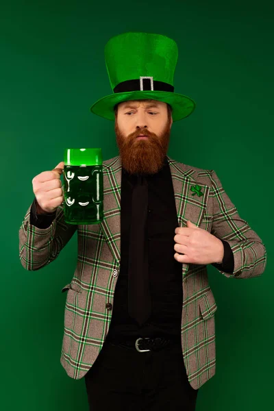 Homem atencioso de chapéu e casaco xadrez segurando vidro de cerveja isolado em verde — Fotografia de Stock