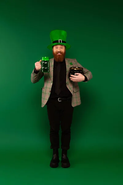 Happy bearded man in hat with clover holding glass of beer and pot with coins on green background — Photo de stock