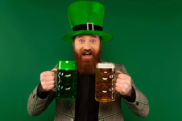 Homem positivo no chapéu segurando copos de cerveja durante a celebração do dia de São Patrício isolado no verde — Fotografia de Stock