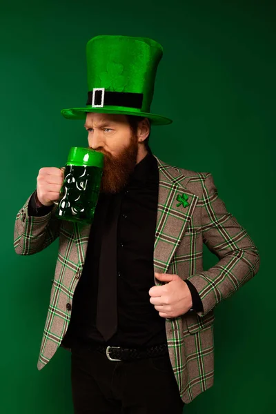 Serious man in hat with clover drinking beer during saint patrick day isolated on green — Stockfoto