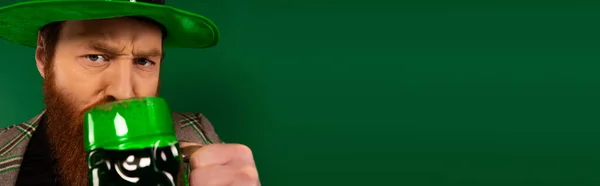 Hombre serio en sombrero bebiendo cerveza mientras celebra el día de San Patricio aislado en verde, pancarta - foto de stock