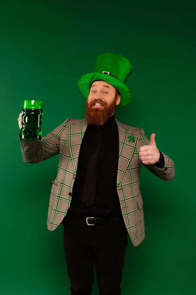 Sorrindo barbudo homem de chapéu segurando cerveja e mostrando como isolado em verde — Fotografia de Stock