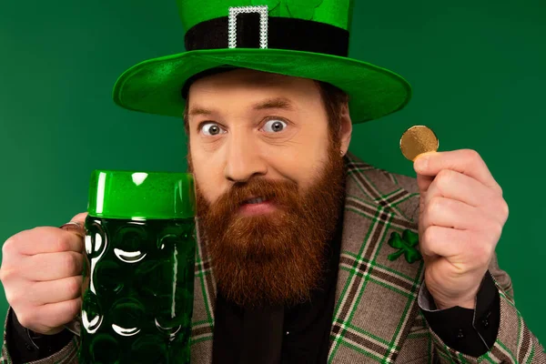 Homem confuso em chapéu segurando moeda de ouro e copo de cerveja isolado em verde — Fotografia de Stock